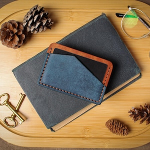 May include: A blue and brown leather cardholder rests on a closed book. The cardholder has a blue front pocket and brown trim. A pair of gold-rimmed glasses, two pinecones, and a set of gold keys are also on the wooden tray.