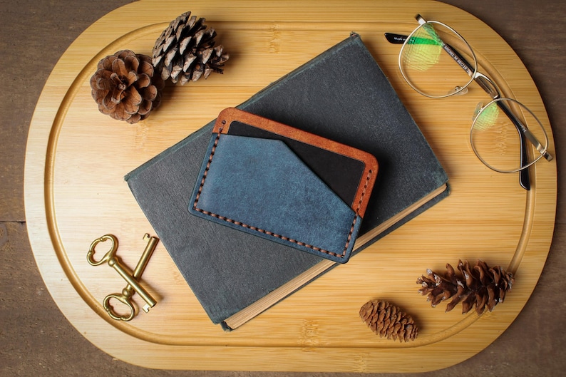May include: A blue and brown leather cardholder rests on a closed book. The cardholder has a blue front pocket and brown trim. A pair of gold-rimmed glasses, two pinecones, and a set of gold keys are also on the wooden tray.