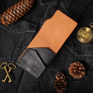 Puede incluir: Una cartera de cuero negro con interior marr&oacute;n, exhibida sobre una chaqueta vaquera negra. La cartera est&aacute; junto a un reloj de bolsillo dorado, dos llaves doradas y pi&ntilde;as. La cartera tiene un borde cosido y un peque&ntilde;o logotipo en relieve.