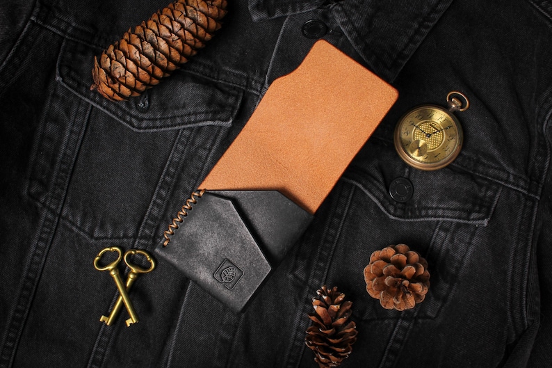 Puede incluir: Una cartera de cuero negro con interior marr&oacute;n, exhibida sobre una chaqueta vaquera negra. La cartera est&aacute; junto a un reloj de bolsillo dorado, dos llaves doradas y pi&ntilde;as. La cartera tiene un borde cosido y un peque&ntilde;o logotipo en relieve.