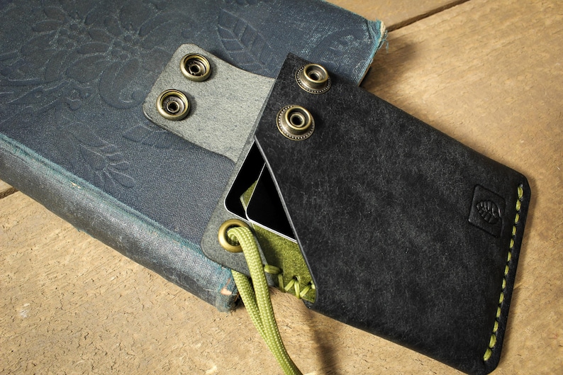 May include: A black leather wallet with a green stitching detail and a small leaf logo. The wallet is attached to a green cord and has a flap with three brass snaps.
