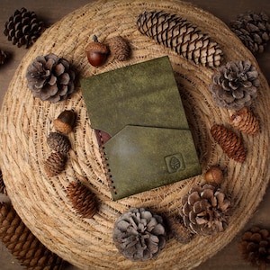 May include: A rectangular olive green leather wallet featuring a diagonal front pocket and a small embossed leaf logo in the bottom right corner. The wallet has visible dark brown stitching along the side edge and a smooth, matte finish.