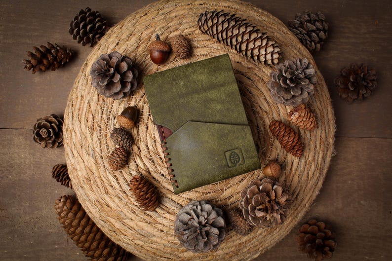 May include: A rectangular olive green leather wallet featuring a diagonal front pocket and a small embossed leaf logo in the bottom right corner. The wallet has visible dark brown stitching along the side edge and a smooth, matte finish.