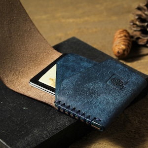 May include: A blue leather wallet with a brown leather flap and a small card slot. The wallet has a stitched edge and a small logo on the front.