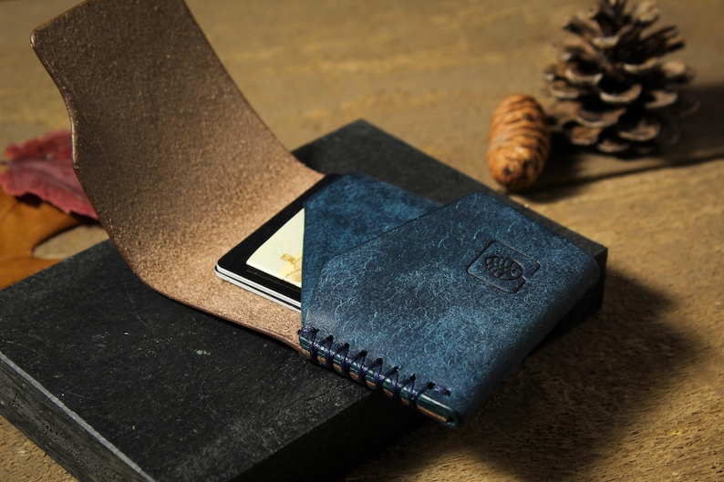 May include: A blue leather wallet with a brown leather flap and a small card slot. The wallet has a stitched edge and a small logo on the front.