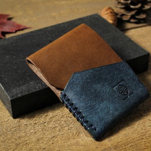 Puede incluir: Cartera de cuero marr&oacute;n y azul de dos pliegues, con borde cosido y emblema en relieve. La cartera descansa sobre un bloque gris oscuro, con hojas de oto&ntilde;o y una pi&ntilde;a en el fondo.