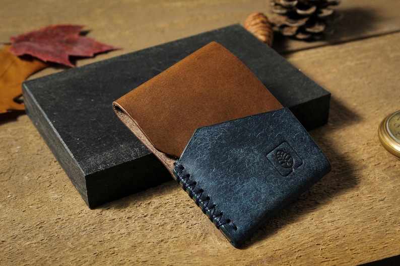 Puede incluir: Cartera de cuero marr&oacute;n y azul de dos pliegues, con borde cosido y emblema en relieve. La cartera descansa sobre un bloque gris oscuro, con hojas de oto&ntilde;o y una pi&ntilde;a en el fondo.