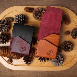 Puede incluir: Vista superior de tres carteras de cuero en negro, burdeos y marr&oacute;n claro, dispuestas sobre una bandeja de madera con pi&ntilde;as. Las carteras tienen un dise&ntilde;o minimalista con un logotipo de &aacute;rbol. El fondo es una superficie de madera.