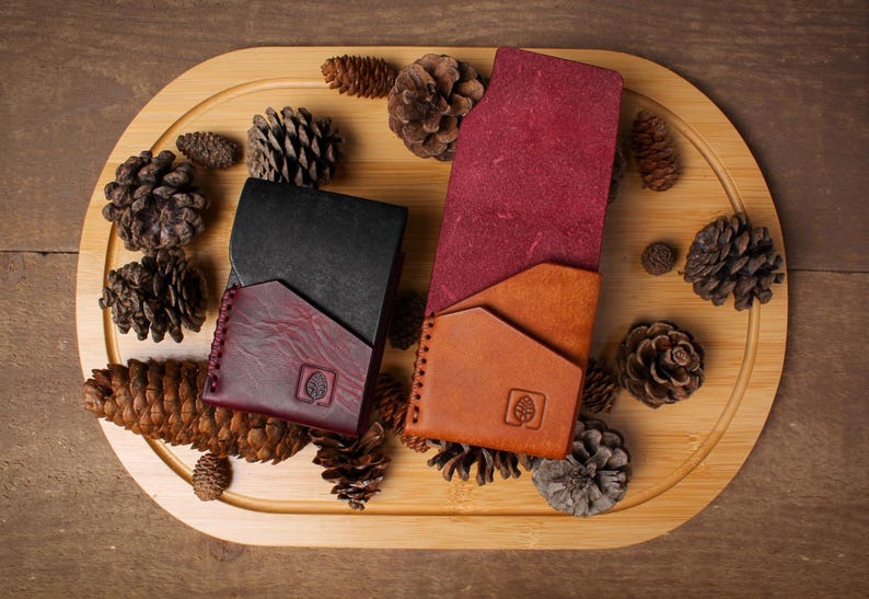 Puede incluir: Vista superior de tres carteras de cuero en negro, burdeos y marr&oacute;n claro, dispuestas sobre una bandeja de madera con pi&ntilde;as. Las carteras tienen un dise&ntilde;o minimalista con un logotipo de &aacute;rbol. El fondo es una superficie de madera.