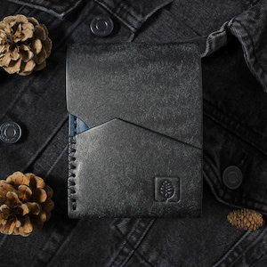 May include: A dark charcoal gray leather wallet featuring a geometric angled pocket design, visible heavy-duty stitching along the left side, and a small embossed square logo depicting a pinecone on the bottom right corner of the front surface.