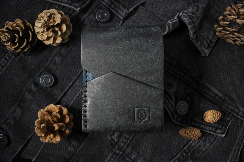 May include: A dark charcoal gray leather wallet featuring a geometric angled pocket design, visible heavy-duty stitching along the left side, and a small embossed square logo depicting a pinecone on the bottom right corner of the front surface.