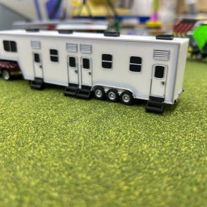 May include: White toy trailer with multiple windows and doors, attached to a red pickup truck. The trailer is on a green grassy surface.