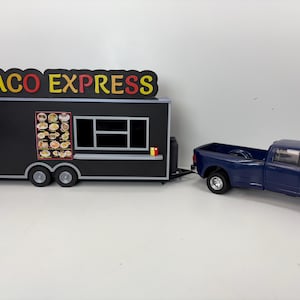 Könnte beinhalten: Ein Miniatur-Taco-Express-Imbisswagen in Schwarz mit einer Menüanzeige und einem Fenster, der an einen blauen Pickup-Truck angehängt ist. Das Schild oben auf dem LKW hat rote und gelbe Buchstaben. Der LKW hat zwei Achsen.