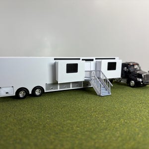 May include: A scale model of a black semi-truck pulling a white trailer with slide-out sections. The trailer features a white exterior, black-framed windows, and a set of silver stairs leading to a central door. The truck has a black cab and chassis.