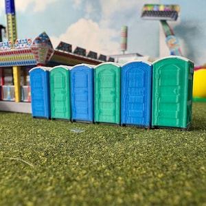 Può includere: Una fila di bagni portatili in miniatura nei toni del blu e del verde è disposta su una superficie verde. Lo sfondo presenta una scena di carnevale con giostre e un cielo nuvoloso, che suggerisce una fiera o un parco divertimenti.