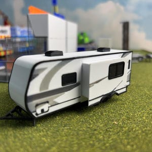 Può includere: Un camper giocattolo bianco e grigio con dettagli neri. Il camper in miniatura ha due piccole finestre e due prese d'aria nere sul tetto. Il camper è su una superficie verde.