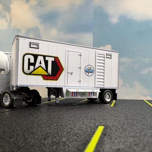 May include: A white trailer with the CAT logo in black, yellow, and red. The trailer has a door, windows, and a small graphic on the side. The trailer is on a black surface with yellow lines, and the background is a blue sky with clouds.