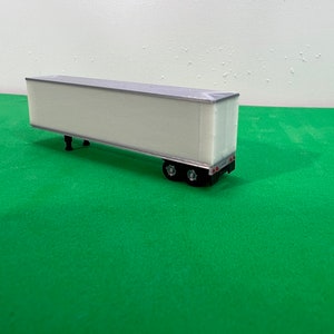 Puede incluir: Un modelo de camión de remolque blanco con ruedas negras y luces traseras rojas. El modelo está sentado sobre una superficie verde.