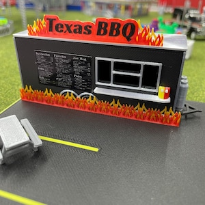 Puede incluir: Un modelo en miniatura de un camión de comida con un exterior negro y un letrero rojo que dice "Texas BBQ". El camión tiene una ventana con un menú y una pequeña mesa y sillas en frente.