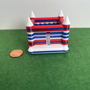 May include: A small red, white, and blue inflatable bounce house with a net-like opening in the front. The bounce house is on a green surface.