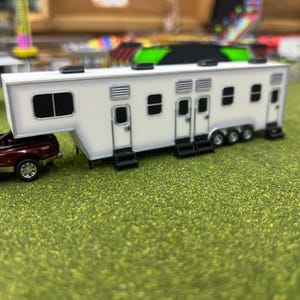 May include: A white toy travel trailer with multiple doors and windows, pulled by a red pickup truck. The trailer is on a green grassy surface.