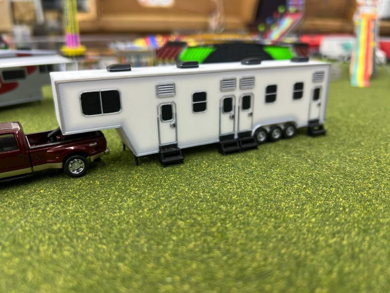 May include: A white toy travel trailer with multiple doors and windows, pulled by a red pickup truck. The trailer is on a green grassy surface.