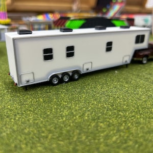 May include: White toy trailer with black accents and three windows. The trailer is attached to a smaller, dark red toy truck.