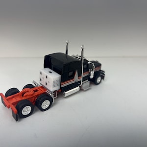 May include: Black and orange semi-truck toy model with a white box on the back. The truck has chrome accents and a silver grill.