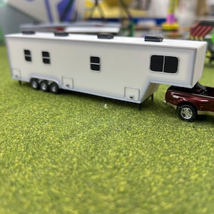 May include: A white toy trailer with black windows is being towed by a burgundy pickup truck. The truck and trailer are on a green grassy surface.