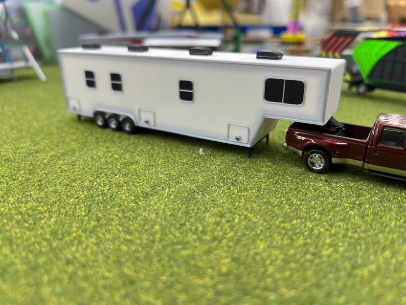 May include: A white toy trailer with black windows is being towed by a burgundy pickup truck. The truck and trailer are on a green grassy surface.