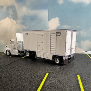 May include: A white semi-truck and trailer model. The trailer has a rectangular shape with a door, windows, and a ladder. The truck is connected to the trailer. The model is on a black surface with yellow lines, against a sky backdrop.