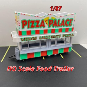 May include: A white and green HO scale food trailer with a red and white striped awning. The awning has the words "Pizza Palace" in red and green letters. The trailer has a sign that says "Wings Garlic Knots Pizza".