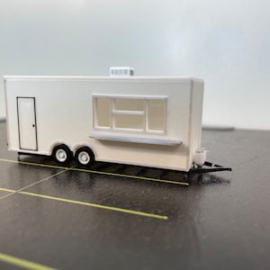 May include: White food truck model with a black door, a window with a counter, and black wheels. The truck is on a black surface.