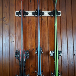 May include: Three pairs of skis are mounted on a wooden wall rack. The skis are black, blue, and green with black bindings.