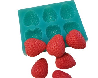 May include: A teal silicone mold with six strawberry-shaped cavities, alongside six red strawberries. The strawberries have a textured surface, and the mold is on a white background. The image showcases a food-themed product.
