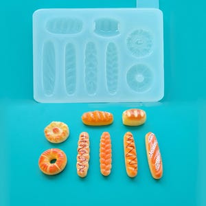 May include: A light blue silicone mold with various bread shapes, including baguettes, rolls, and a donut. Several miniature bread replicas, painted in shades of brown and orange, are arranged below the mold on a teal background. The image showcases a baking-themed craft.