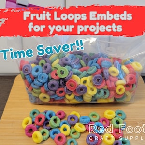 May include: A clear plastic bag filled with colorful Fruit Loops cereal. The bag is labeled "Fruit Loops Embeds for your projects" and "Time Saver!!". A small pile of Fruit Loops is on a wooden surface below the bag. The text "Red Foot Craft Supply" is visible below the pile of cereal.