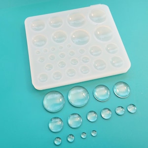 May include: A clear, flexible silicone mold with various sized dome shapes, alongside several clear, domed resin pieces. The mold is white and square, with rounded edges. The resin pieces are arranged on a teal surface.