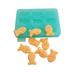 May include: A teal silicone mold with fish-shaped cavities, partially filled with yellow fish-shaped crackers. Several crackers are scattered below the mold. The crackers are a light orange color.