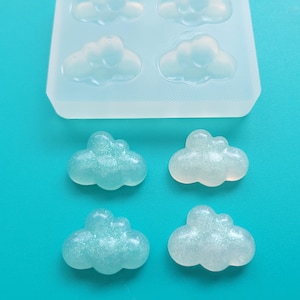 May include: A clear silicone mold with cloud-shaped cavities, alongside four cloud-shaped resin pieces with glitter. The clouds are light blue and white, set against a bright teal background. The image showcases a craft project.