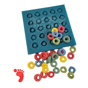 May include: A teal silicone mold with circular cavities, partially filled with colorful, ring-shaped craft supplies. Scattered around the mold are additional rings in red, yellow, green, blue, and other colors. The image includes the text "Red Foot CRAFT SUPPLY".