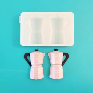 May include: A white silicone mold featuring two cavities shaped like moka pots, positioned above two finished white moka pot figures with black handles. The figures have a faceted design and a distinct central band, mirroring the shape of the mold.