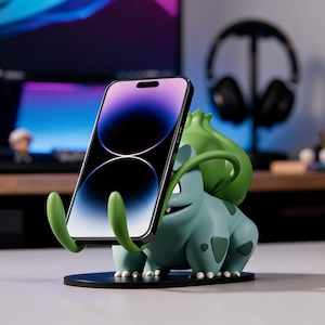 May include: A phone stand featuring a blue and green Bulbasaur figure holding a smartphone. The stand has a black base and green arms to cradle the phone. The smartphone screen displays a dark background.