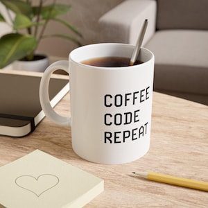 Peut inclure: Une tasse en céramique blanche avec une anse, remplie de café brun foncé et une cuillère en argent. Le texte COFFEE CODE REPEAT est imprimé en lettres capitales noires, grasses et sans empattement sur le devant, disposé sur trois lignes.