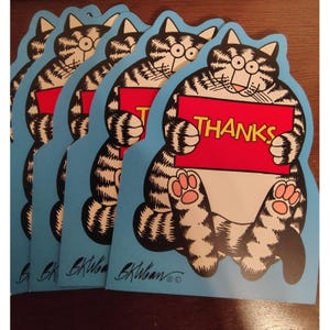 May include: A stack of blue cat-shaped greeting cards. Each card features a cartoon cat holding a red sign with the word "THANKS" in white. The cat is black and white with pink paw pads. The artist's signature is at the bottom.