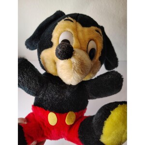 May include: A vintage plush toy of a dog-like character, predominantly black and yellow, with large eyes and a black nose. The toy has black ears, arms, and legs, with red trousers and yellow buttons. The plush is in a sitting position with arms outstretched.