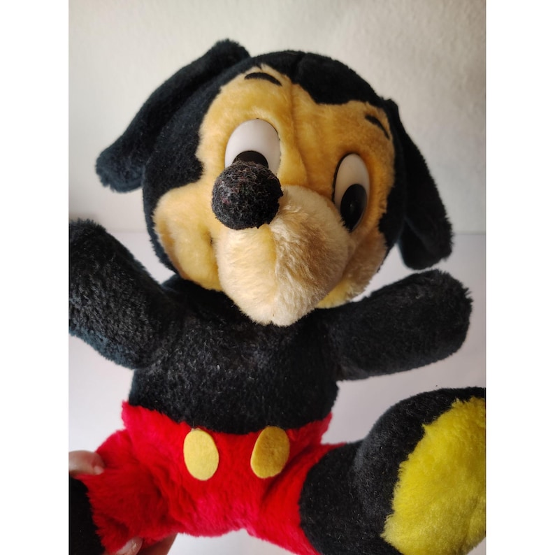May include: A vintage plush toy of a dog-like character, predominantly black and yellow, with large eyes and a black nose. The toy has black ears, arms, and legs, with red trousers and yellow buttons. The plush is in a sitting position with arms outstretched.