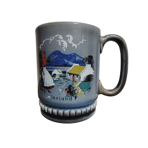 Puede incluir: Taza de cerámica gris con asa y base decorativa. La taza presenta una ilustración de un paisaje irlandés con una mujer con vestimenta tradicional, casas de campo y montañas. La palabra "Ireland" está impresa en la taza.