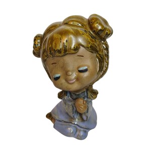 Pode incluir: Uma figura de cerâmica de uma menina com cabelo castanho e um vestido roxo. Ela está ajoelhada com as mãos juntas em oração.