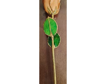May include: A single preserved rose with a peach-colored bloom and gold-trimmed green leaves. The stem is gold-colored. The rose is displayed against a dark wood background.